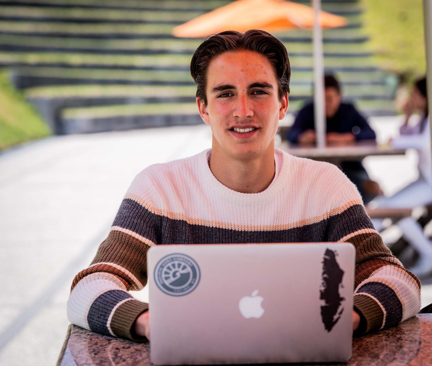 Joven estudiante sonriendo a la cámara mientras usa su laptop en una mesa al aire libre en el campus.